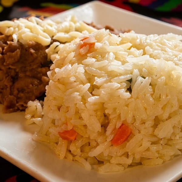 A white plate holds a serving of fluffy white rice with vegetables and refried beans topped with cheese.